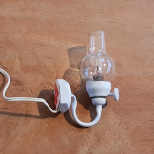 Op de afbeelding: Een witte wandlamp met een heldere glazen bolkap en een kleine instelknop. Het armatuur heeft een gebogen witte metalen arm, een witte voet met een rood accent en een wit elektrisch snoer dat uit de zijkant van de voet komt.