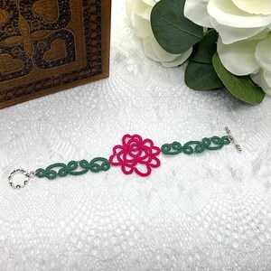 May include: A green and pink crocheted bracelet with a rose design and a silver toggle clasp.