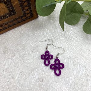 May include: A pair of purple crocheted cross earrings with silver hooks. The earrings are on a white lace background.