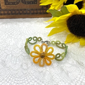 May include: A handmade, delicate bracelet featuring a central flower design and a series of connected loops. The flower is yellow and orange with a brown center. The bracelet is green and yellow, with a silver clasp. The bracelet is displayed on a white lace surface.