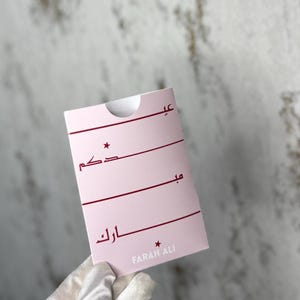 May include: A pink card with burgundy text and a star design. The card has Arabic script and the name "FARAH ALI" at the bottom. The card is held by a gloved hand.