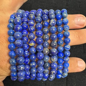 Puede incluir: Múltiples pulseras de cuentas de lapislázuli azul. Las pulseras están hechas de cuentas redondas y pulidas en varios tonos de azul con motas doradas. Las pulseras están apiladas, mostrando el color y la textura de las piedras preciosas.
