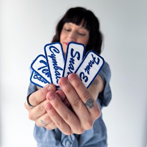 Op de afbeelding: Een persoon houdt vijf witte rechthoekige patches vast met blauw geborduurde tekst. De tekst op de patches luidt "Ten Bass", "Cymbals", "Snare" en "Front B". De patches hebben een blauwe rand.