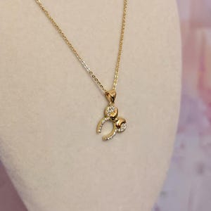 Puede incluir: Un collar dorado con un colgante con forma de figura estilizada. El colgante presenta piedras claras y brillantes y está suspendido de una delicada cadena. El collar se muestra sobre un fondo suave y neutro.