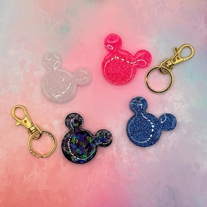 Puede incluir: Cuatro llaveros con forma de ratón hechos de resina con purpurina. Uno es blanco translúcido, uno rosa brillante, uno azul oscuro y uno negro con confeti iridiscente. Se incluyen dos cierres de mosquetón de metal dorado con anillas.