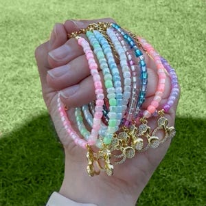 Puede incluir: Una colección de pulseras de cuentas coloridas en tonos rosa, verde, azul y morado. Cada pulsera presenta un dije dorado con un diseño brillante. Las pulseras están unidas por una cadena dorada.