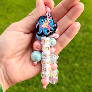 May include: A keychain featuring a rubber charm of Eeyore and Piglet hugging, suspended above a vertical column of white square beads spelling EEYORE in gold letters, surrounded by various round beads in shades of pink, blue, and iridescent white.