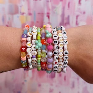 Puede incluir: Una colección de pulseras de cuentas coloridas apiladas en una muñeca. Las pulseras presentan varios colores, incluyendo rosa, verde y blanco, con cuentas con letras que deletrean palabras como "HEAL", "PERDITA" y "GUESTKIN".