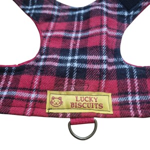 May include: A red and black plaid dog harness featuring a yellow label with the text "LUCKY BISCUITS". The harness includes a D-ring for a lead and a black Velcro fastening. The plaid design incorporates white lines.