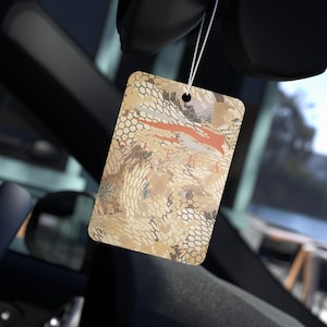 May include: A beige and brown car air freshener with a camouflage pattern and a red stripe. The air freshener is hanging from a rearview mirror.