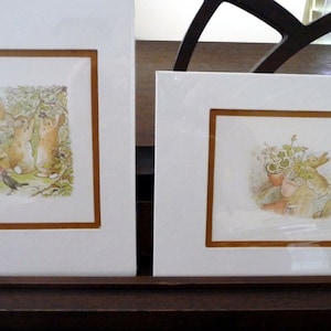 May include: Two framed illustrations of rabbits. The first illustration shows two rabbits standing on their hind legs, with a black bird in the foreground. The second illustration shows a rabbit sitting in a pot of flowers.