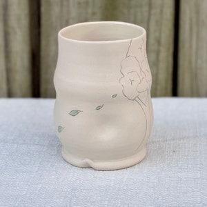 May include: A white ceramic mug with a hand-drawn design of a tree with leaves blowing in the wind. The mug has a rounded shape and a slightly uneven base.