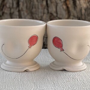 May include: Two white ceramic mugs with a red balloon design. The balloons are drawn with black lines and have a red fill. The mugs have a rounded shape and a wide base.