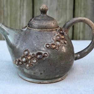 May include: A gray ceramic teapot with a lid and a handle. The teapot has a textured surface with small, raised circles. The teapot is sitting on a white surface.