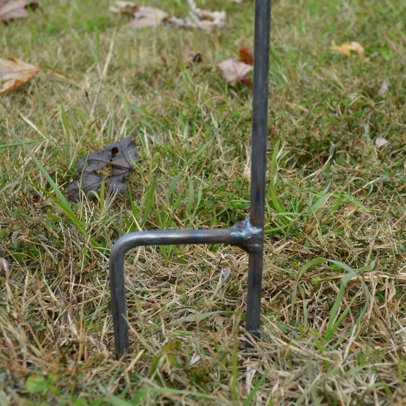 Op de afbeelding: Close-up van een metalen paal met een gebogen arm, ontworpen om in de grond te worden gestoken. De paal is gemaakt van donkergrijs metaal en is gedeeltelijk in de grond gestoken, die bedekt is met bruin gras en bladeren.