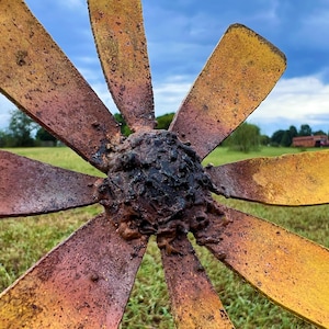 May include: Close-up of a weathered metal sculpture resembling a stylized flower or fan. The blades are a mix of yellow and rust-colored hues, radiating from a dark, textured center. The background shows a green field and a cloudy sky.
