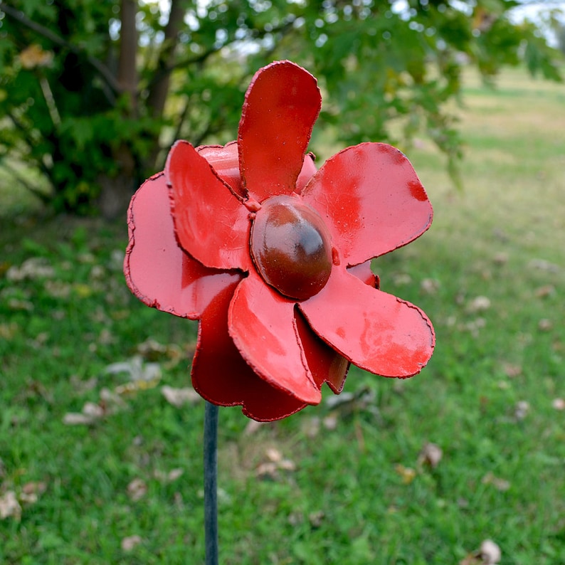 Puede incluir: Una flor de metal roja con seis p&eacute;talos y un centro marr&oacute;n. La flor est&aacute; unida a un tallo de metal.