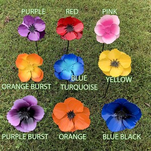 Op de afbeelding: Negen metalen bloemen in verschillende kleuren, waaronder paars, rood, roze, oranje, blauw, turquoise, geel en zwart. De bloemen zijn in een cirkel op een groene grasmat gerangschikt.