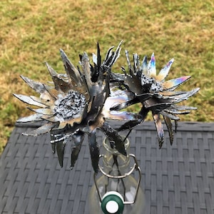 Op de afbeelding: Drie metalen bloemen in een heldere glazen fles. De bloemen zijn gemaakt van metaal en hebben een donkere, iriserende afwerking. De bloemen zijn in een vaas gerangschikt en klaar om te worden tentoongesteld.