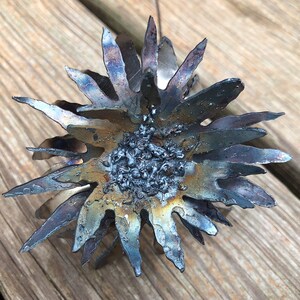 Op de afbeelding: Een metalen zonnebloem sculptuur met een getextureerde, gebrande afwerking. De bloemblaadjes zijn dun en puntig, en het midden van de bloem is gevuld met kleine, ronde, metalen kralen.