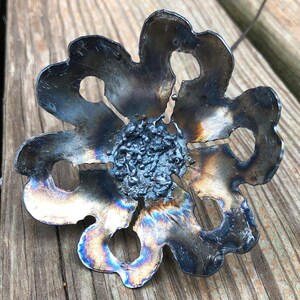 K&ouml;nnte beinhalten: Eine Metallblume mit dunkelblauer und irisierender Oberfl&auml;che. Die Blume hat eine Mitte mit einer strukturierten, dunkelgrauen Oberfl&auml;che.