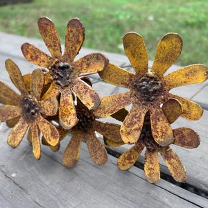 May include: A cluster of four metal daisy flowers with a rustic, weathered appearance. The petals are yellow with brown rust spots, and the centers are dark brown. The flowers are arranged on a wooden surface.