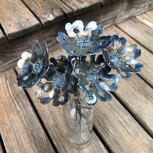 K&ouml;nnte beinhalten: Ein Strau&szlig; aus Metallblumen in einer durchsichtigen Glasflasche. Die Blumen sind aus Metall und haben eine dunkelgraue Oberfl&auml;che. Die Blumen sind in einer Vase arrangiert und bereit zum Ausstellen.