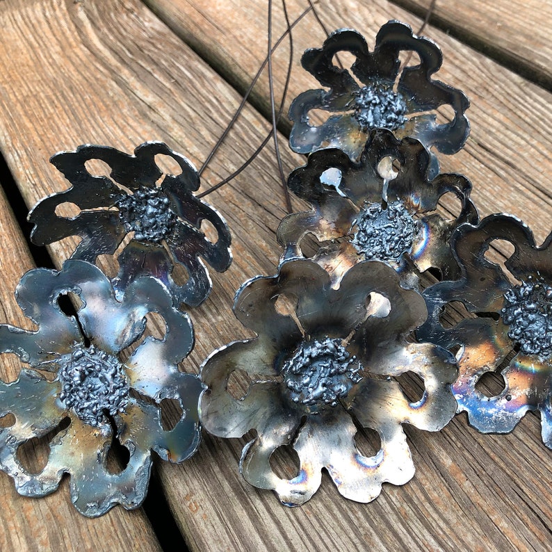 K&ouml;nnte beinhalten: Ein Strau&szlig; aus sieben Metallblumen mit rustikalem, gealtertem Finish. Jede Blume hat eine einzigartige Form und Textur, mit einer Mitte, die einer kleinen, strukturierten Kugel &auml;hnelt. Die Blumen sind an d&uuml;nnen Metallstielen befestigt.