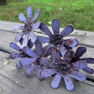 May include: A cluster of metal daisy flowers with a distressed, rustic appearance. The petals are a muted purple with rusty brown accents, and the flowers are arranged on a wooden surface. The overall aesthetic is vintage and handcrafted.