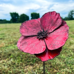 Pu&ograve; includere: Una grande scultura floreale in metallo realizzata a mano, di colore rosso intenso e con un centro scuro. I petali hanno un aspetto strutturato e la scultura &egrave; montata su un sottile stelo di metallo nero, su un campo erboso.