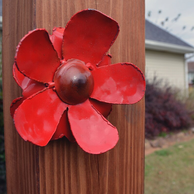 Puede incluir: Una flor de metal roja con cinco p&eacute;talos est&aacute; unida a un poste de madera. La flor tiene un centro marr&oacute;n.