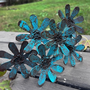 May include: A cluster of six metal flower sculptures with a distressed turquoise finish and rusty brown accents. The flowers are arranged on a weathered wooden surface, with a blurred green background.