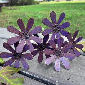 May include: A cluster of handcrafted metal flowers with purple petals and dark centers. The flowers are arranged on a wooden surface with green foliage in the background. The metal has a rustic, weathered appearance.