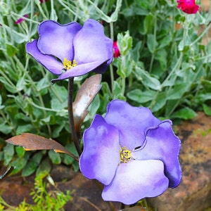 May include: Two purple metal flowers with yellow centers. The flowers are on a rusty metal stem and are surrounded by green foliage.