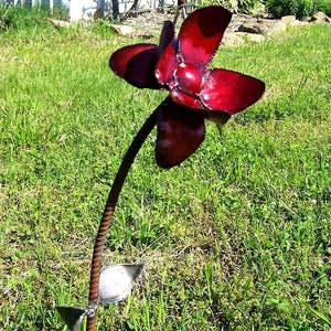 Op de afbeelding: Een rode metalen bloemsculptuur met een lange stengel en bladeren. De bloem is gemaakt van meerdere bloemblaadjes en heeft een glanzende afwerking.