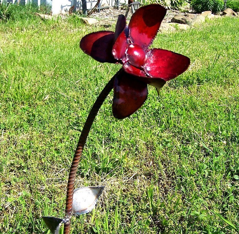 Op de afbeelding: Een rode metalen bloemsculptuur met een lange stengel en bladeren. De bloem is gemaakt van meerdere bloemblaadjes en heeft een glanzende afwerking.