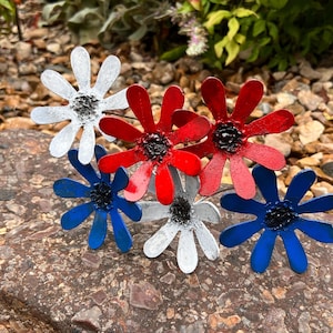 May include: A collection of metal flower sculptures in red, white, and blue. The flowers have a daisy-like design with black centers. They are arranged on a brown rock, with greenery in the background. The flowers are likely garden decor.