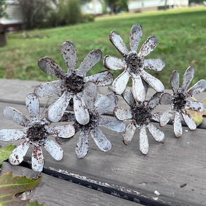 May include: A cluster of six metal flower sculptures with a distressed, aged appearance. The flowers have white petals with brown rust spots and dark centers. The arrangement sits on a weathered wooden surface.