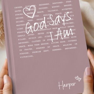 May include: A rosewood-colored journal with the words "God says I Am" in white script. The journal has a heart graphic and the name "Harper" at the bottom. The journal is held in two hands.