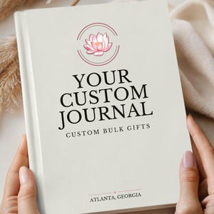 May include: Ivory-colored journal with the text "YOUR CUSTOM JOURNAL" and a lotus flower logo. The journal is held open, displaying the text "CUSTOM BULK GIFTS" and "ATLANTA, GEORGIA". The journal is a custom bulk gift.