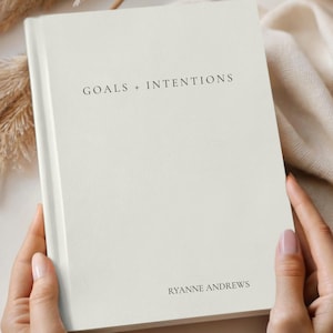 May include: Ivory-colored journal with the text "GOALS + INTENTIONS" and "RYANNE ANDREWS" printed on the cover. The journal is held by hands and is surrounded by neutral-toned decor. The journal is a product of Comfort & Joy Co.