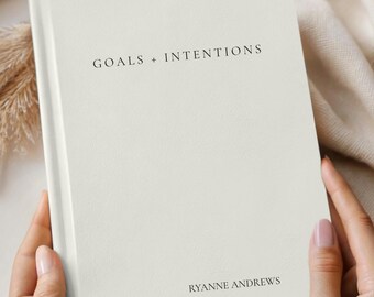May include: Ivory journal with the text "GOALS + INTENTIONS" and "RYANNE ANDREWS" printed on the cover. The journal is held by hands and is surrounded by neutral-toned decor. The journal is a product of Comfort & Joy Co.