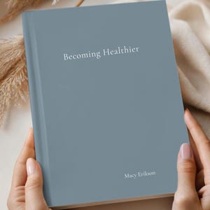 May include: A steel-colored journal titled "Becoming Healthier" with the author's name, Macy Erikson, printed on the cover. The journal is held by a person. The Comfort & Joy Co. logo is in the lower left corner.