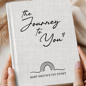 May include: A book with a faux linen ivory cover titled "The Journey to You" in black script. The cover features a rainbow graphic and the text "Baby Smith's IVF Story". The book is held by a person.