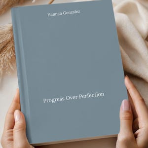 May include: A book with a steel-colored cover held by hands. The book's title is "Progress Over Perfection" and the author's name is "Hannah Gonzalez". The top of the book has the word "STEEL" printed on it.