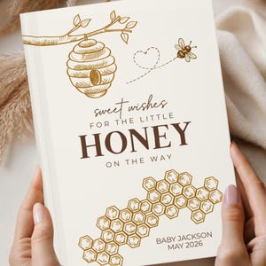 May include: A cream-colored baby announcement book with a bee and honeycomb design. The text reads "sweet wishes for the little HONEY on the way." The book is held open, revealing the text and illustrations. The baby's name and due date are also printed on the book.