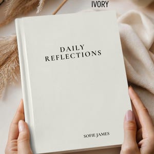 May include: An ivory-colored journal titled "Daily Reflections" by Sofie James. The book is held open by a person, showcasing the cover's minimalist design. The top of the journal is labeled "IVORY". The journal is surrounded by neutral-toned decor.
