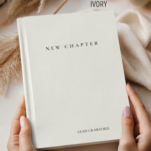 May include: Ivory-colored book with the words "NEW CHAPTER" and "LEAH CRAWFORD" printed in black. The book is held by a person. The words "Comfort & Joy Co." are in the lower left corner. The book is on a white surface.