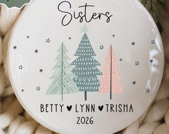 May include: A round white ceramic ornament featuring the text Sisters at the top, three stylized Christmas trees in light green, blue, and pink, and the names Betty, Lynn, and Trisha separated by hearts, with the year 2026 printed below.