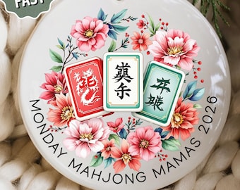 May include: A round white ceramic ornament featuring a central illustration of three mahjong tiles surrounded by pink and red flowers. The text MONDAY MAHJONG MAMAS 2026 is printed in a circular arc along the bottom edge of the ornament.